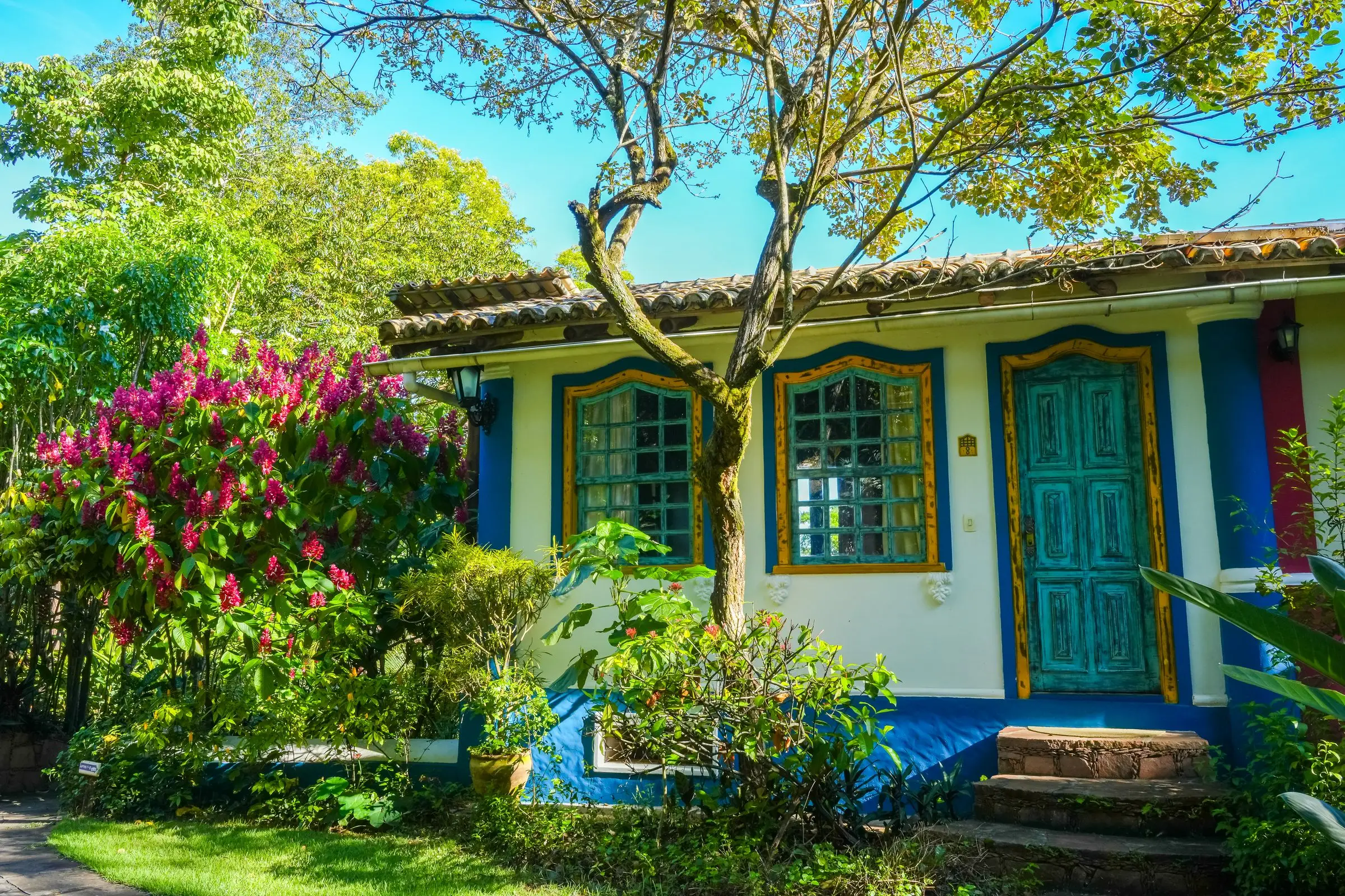 Fachada azul e branca com portas de madeira entre flores e árvores do jardim