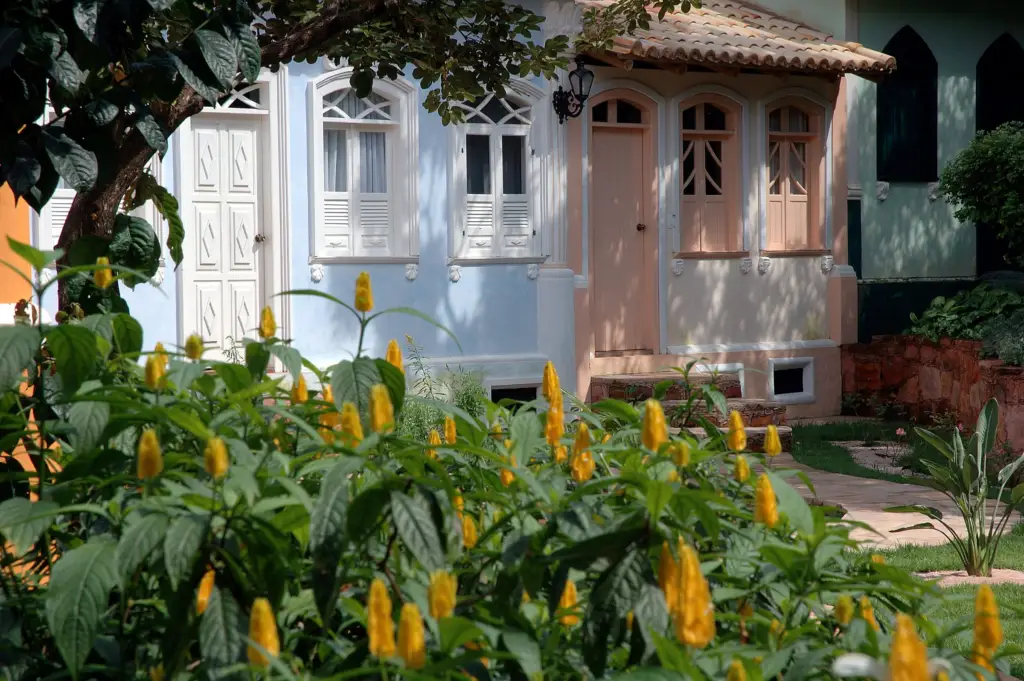 Casarão azul-claro de janelas brancas atrás de flores amarelas no jardim da Vila Serrano